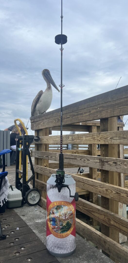 A pelican perched on a pier railing next to a fishing rod and a propane tank with a colorful cover.