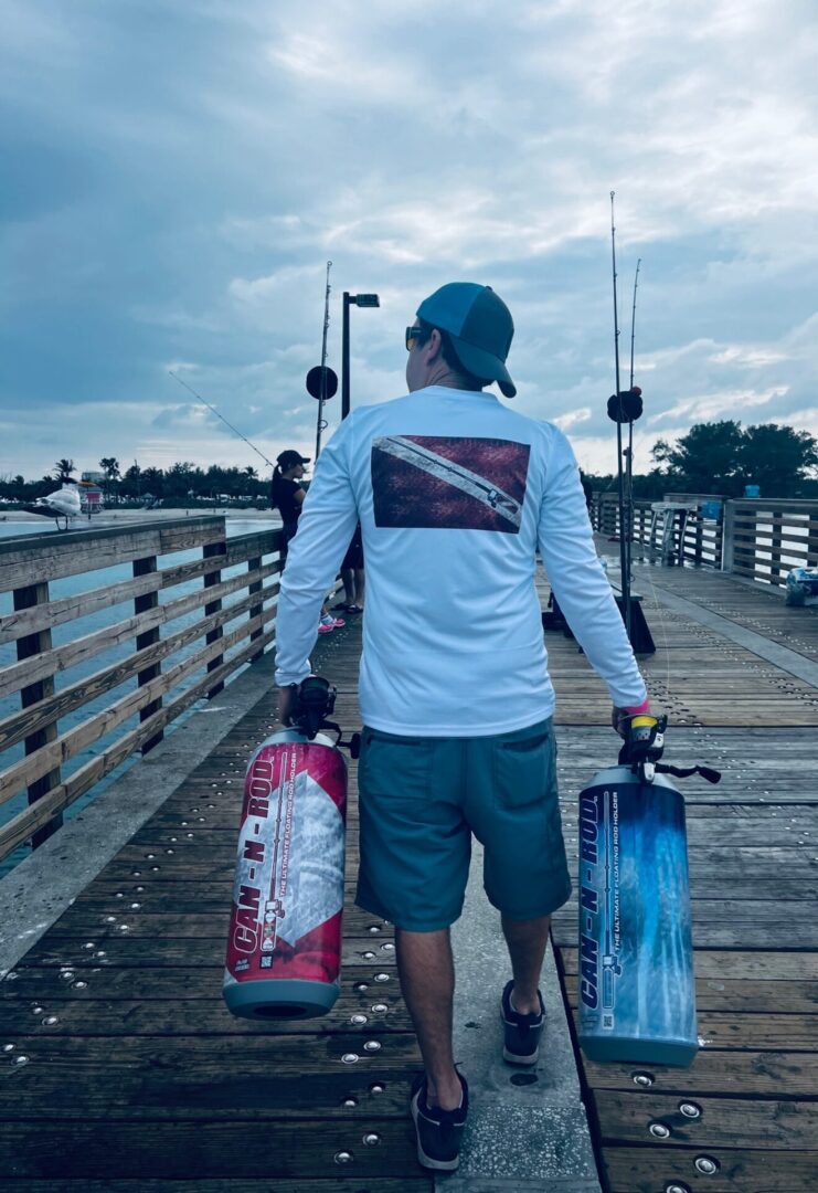 Man carrying two fishing rods and portable coolers walking on a pier, with overcast skies and other fishermen in the background.