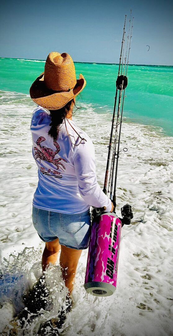 Woman in a straw hat and white shirt wading into turquoise water, carrying a fishing rod and a pink tackle box.