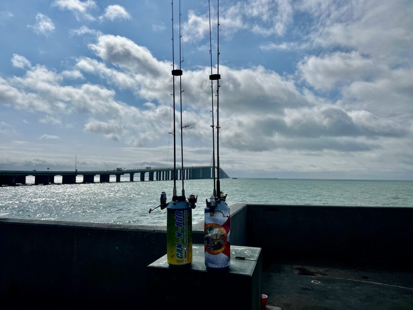 Two fishing rods leaning against a wall with bait containers, overlooking a calm sea and a distant pier under a partly cloudy sky.